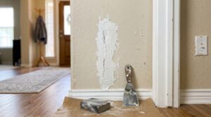 A textured interior wall with freshly applied white joint compound over a vertical crack next to a putty knife and sanding block, demonstrating essential drywall repair tips.